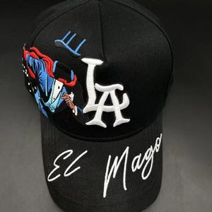 K&ouml;nnte beinhalten: Schwarze Baseballkappe mit wei&szlig; gestickten "LA"-Buchstaben und den Worten "El Mago" in Schreibschrift. Die Kappe hat ein farbenfrohes Stickdesign eines Zauberers auf der Seite.