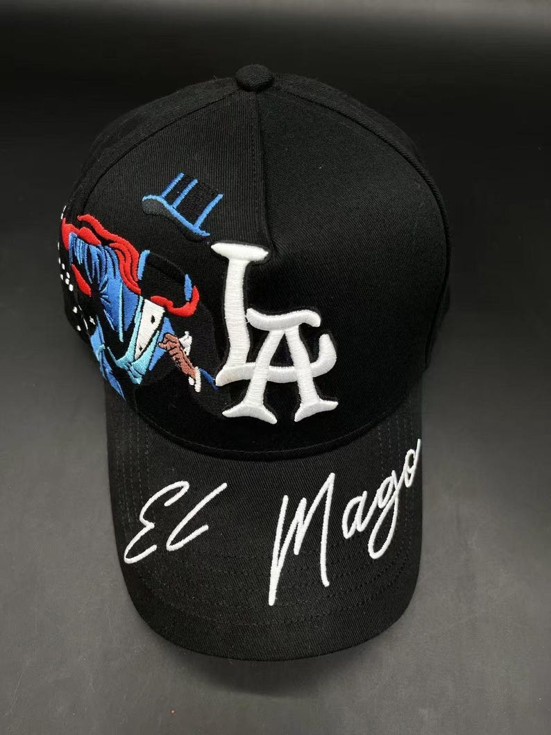 K&ouml;nnte beinhalten: Schwarze Baseballkappe mit wei&szlig; gestickten "LA"-Buchstaben und den Worten "El Mago" in Schreibschrift. Die Kappe hat ein farbenfrohes Stickdesign eines Zauberers auf der Seite.