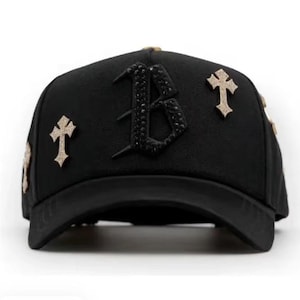 May include: Black baseball cap with a curved brim. The cap features a large, jeweled letter "B" on the front and several cross-shaped embellishments. The cap is a solid black color with gold-colored accents.