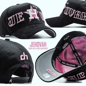 May include: Black baseball cap with the word "JEHOVAH" embroidered in pink. The hat features a pink star and lightning bolt design. The underside of the brim is gray, and the interior is pink. The text "DANDY HATS X RUDE AWAKENINGS" is also visible.