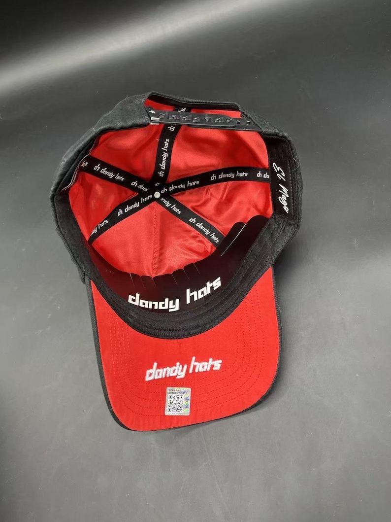 K&ouml;nnte beinhalten: Schwarze und rote Baseballkappe mit dem Text "dandy hats" auf dem Schirm und innen. Die Kappe hat einen Snapback-Verschluss und den Text "El Mago" auf der Seite. Im Inneren befindet sich der Text "ch dandy hats".