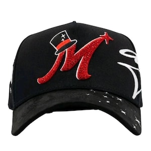 May include: Black baseball cap with a curved brim. The cap features a red glitter "M" with a star and a top hat design. The brim has a pattern of white stars.