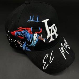 K&ouml;nnte beinhalten: Schwarze Baseballkappe mit wei&szlig; gesticktem "LA"-Logo und "El Mago"-Text. Verf&uuml;gt &uuml;ber eine farbenfrohe gestickte Grafik eines Zauberers in Aktion. Die Kappe hat einen gebogenen Schirm und einen Knopf oben.