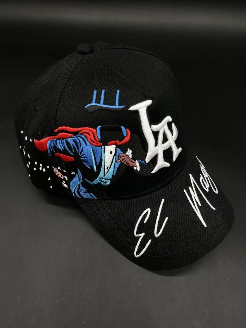 K&ouml;nnte beinhalten: Schwarze Baseballkappe mit wei&szlig; gesticktem "LA"-Logo und "El Mago"-Text. Verf&uuml;gt &uuml;ber eine farbenfrohe gestickte Grafik eines Zauberers in Aktion. Die Kappe hat einen gebogenen Schirm und einen Knopf oben.