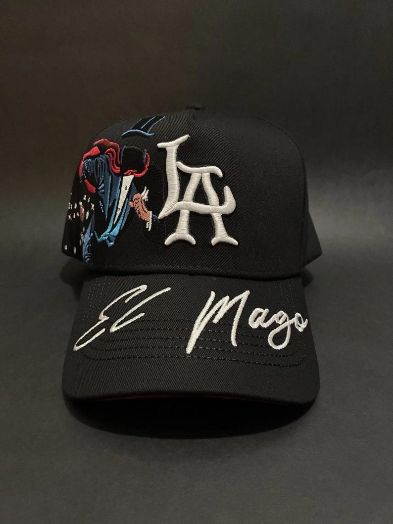 K&ouml;nnte beinhalten: Schwarze Baseballkappe mit wei&szlig; gesticktem "LA"-Logo und "El Mago"-Schriftzug. Die Kappe hat ein farbenfrohes Stickdesign auf der Seite, das diesem stilvollen Accessoire eine einzigartige Note verleiht.