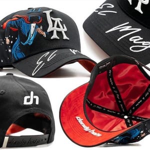 K&ouml;nnte beinhalten: Schwarze Baseballkappe mit wei&szlig;er gestickter Schriftzug "LA" und "El Mago". Verf&uuml;gt &uuml;ber eine farbenfrohe Illustration einer Figur in Anzug und Umhang. Die Kappe hat ein rotes Satinfutter und einen schwarzen verstellbaren Riemen.