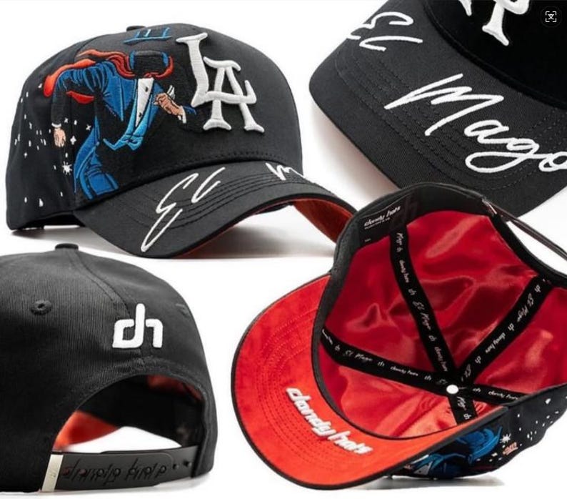 K&ouml;nnte beinhalten: Schwarze Baseballkappe mit wei&szlig;er gestickter Schriftzug "LA" und "El Mago". Verf&uuml;gt &uuml;ber eine farbenfrohe Illustration einer Figur in Anzug und Umhang. Die Kappe hat ein rotes Satinfutter und einen schwarzen verstellbaren Riemen.