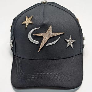 Puede incluir: Gorra de béisbol negra con adornos de estrellas y luna creciente. La gorra tiene una visera curva y un cierre metálico en la parte trasera. Las estrellas son doradas, plateadas y con incrustaciones de cristal.
