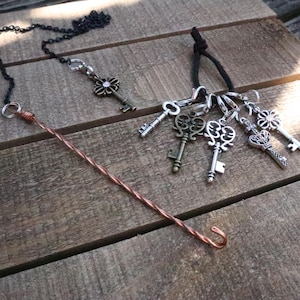 May include: A collection of metal jewelry components including a long twisted copper wire hook, a black chain with a single flower-shaped key charm, and a black cord featuring a cluster of five assorted silver and bronze-toned skeleton key charms.