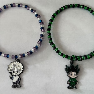 May include: Two beaded bracelets with anime character charms. The bracelet on the left has white and purple beads with a black and white anime character charm. The bracelet on the right has green and black beads with a green and black anime character charm.