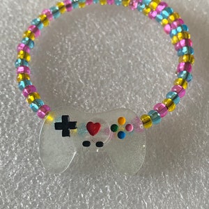 May include: A colorful beaded bracelet with a clear plastic charm shaped like a video game controller. The controller has a red heart, black buttons, and a black plus sign.