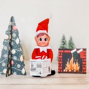 May include: A Christmas elf doll wearing a red suit and hat, holding a newspaper. A decorative Christmas tree and a fireplace scene are on either side of the elf. The newspaper has the word "TECHNOLOGY" printed on it.