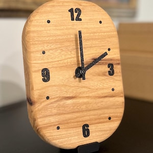 May include: A wooden clock with a light brown finish and black numerals. The clock has a rectangular shape with rounded edges and black hands. The clock is displayed on a black stand.