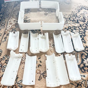 May include: A collection of white plastic components, likely for a modular structure, arranged on a patterned rug. The pieces include curved and rectangular sections, suggesting a building or organizational system.