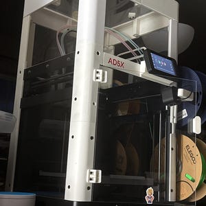 May include: A white and black Flashforge AD5X 3D printer with a clear enclosure. The printer has a small screen and a sticker of a cartoon character. Spools of filament are visible on the side.