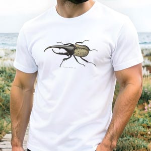 May include: White t-shirt featuring a detailed illustration of a large beetle. The beetle has a dark body with a textured, dotted pattern on its back and prominent horns. The design is centered on the shirt, offering a nature-inspired aesthetic.