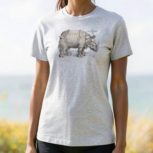 May include: A light gray t-shirt featuring a detailed illustration of a rhinoceros. The rhino is depicted in a vintage style, with intricate shading and lines. The shirt has a classic crew neck and short sleeves, suitable for casual wear.