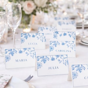 Puede incluir: Decoración de mesa con tarjetas de nombre florales azules y blancas. Los nombres Maria, Julia y Carolina son visibles. Copas de vino y un arreglo floral en el fondo.