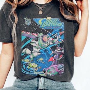 May include: A charcoal gray short-sleeve t-shirt featuring a graphic print of Buzz Lightyear Laser Blast. The design includes Buzz Lightyear, Zurg, and green aliens in space with the text Buzz Lightyear Laser Blast and Discoveryland.