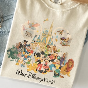May include: Cream-colored t-shirt featuring a colorful graphic of Disney characters and Cinderella's castle. The words "Walt Disney World" are printed in black script at the bottom. The shirt is folded on a pair of blue jeans.