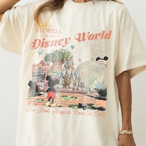 Puede incluir: Camiseta color crema con un dise&ntilde;o gr&aacute;fico de Disney World en Florida. El dise&ntilde;o incluye el texto "Florida Est. 1971 Disney World" y "The Most Magical Place On Earth", con Mickey Mouse, un castillo y una gran esfera.