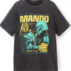 K&ouml;nnte beinhalten: Dunkelgraues T-Shirt mit einer Grafik von Mando und Grogu aus The Mandalorian. Das Design enth&auml;lt den Text "MANDO" in Gelb und "GROGU" in T&uuml;rkis. Der Satz "THE LEGEND CONTINUES" ist ebenfalls vorhanden.