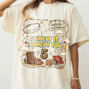 May include: An off-white t-shirt featuring a graphic print with a lasso border, cowboy boots, a red hat, alphabet blocks, and the text "Cinfinity and beyond", "I KNEW IT I KNEW YOU", "5", and "YOU'VE GOT A FRIEND IN ME" with stars and clouds.