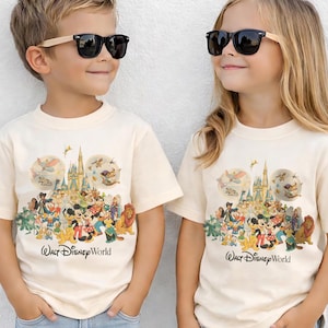 Puede incluir: Dos ni&ntilde;os vistiendo camisetas color crema con un gr&aacute;fico colorido de personajes de Disney y un castillo. Las camisetas tienen el texto "Walt Disney World" impreso. Ambos ni&ntilde;os llevan gafas de sol negras.