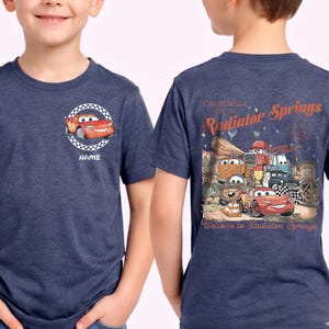 Puede incluir: Camiseta azul marino con un dise&ntilde;o de dibujos animados de Rayo McQueen de la pel&iacute;cula Cars. La parte delantera tiene una imagen circular con la palabra "NAME". La parte trasera presenta un gr&aacute;fico de Radiator Springs, California, con varios personajes de coches.