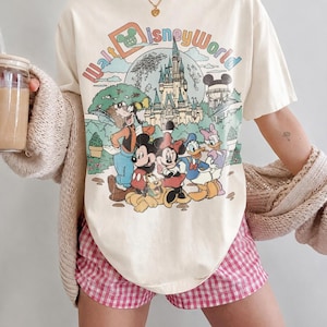May include: Ivory-colored t-shirt featuring a colorful graphic of Disney characters and the text "Walt Disney World." The shirt is paired with pink and white checkered shorts. The words "Comfort Colors" and "Ivory" are printed at the bottom.