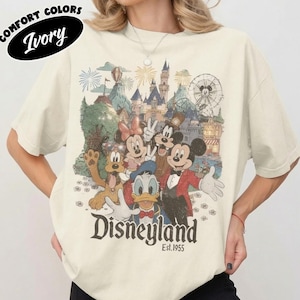 May include: An ivory t-shirt featuring a graphic print of cartoon characters including Mickey Mouse, Minnie Mouse, Donald Duck, Goofy, and Pluto in front of a castle. The text reads Disneyland Est. 1955. The shirt has short sleeves and a crew neckline.
