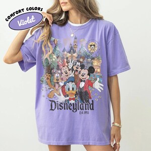 May include: A violet short-sleeve t-shirt featuring a graphic print of Mickey Mouse, Minnie Mouse, Donald Duck, Goofy, and Pluto in front of a castle. The text reads Disneyland Est. 1955. A circular logo in the corner says Comfort Colors Violet.