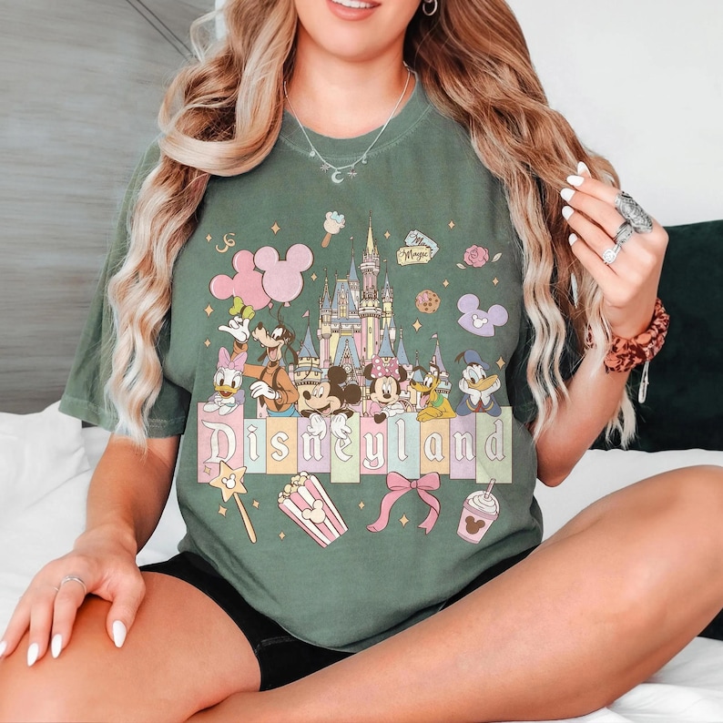 May include: Green t-shirt featuring a Disneyland graphic with cartoon characters, a castle, and the word "Disneyland" in pastel colors. The design includes Mickey Mouse, Minnie Mouse, Donald Duck, and Goofy. The shirt has a relaxed fit.
