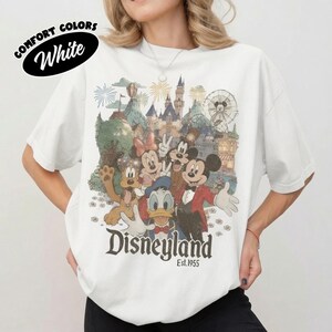 May include: A white t-shirt featuring a vintage-style graphic of Mickey Mouse, Minnie Mouse, Donald Duck, and Pluto in front of a Disneyland castle and Ferris wheel. The text reads Disneyland Est. 1955. A black oval badge says COMFORT COLORS White.