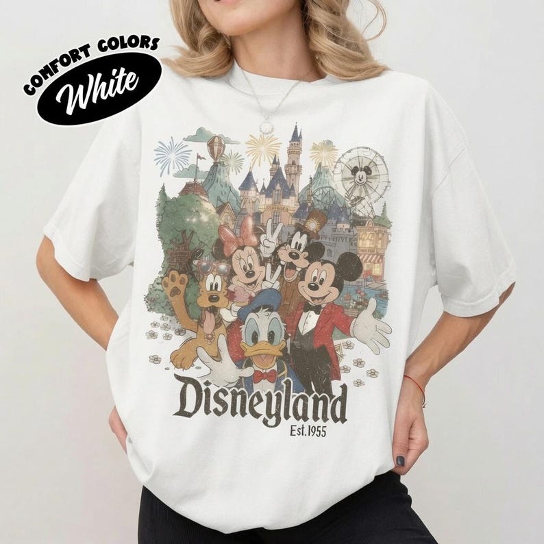 May include: A white t-shirt featuring a vintage-style graphic of Mickey Mouse, Minnie Mouse, Donald Duck, and Pluto in front of a Disneyland castle and Ferris wheel. The text reads Disneyland Est. 1955. A black oval badge says COMFORT COLORS White.