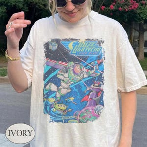 May include: An ivory t-shirt featuring a graphic print of Buzz Lightyear from the Laser Blast attraction. The design includes space imagery, green aliens, and the text Buzz Lightyear Laser Blast and Discoveryland in a rectangular frame.