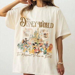 May include: Cream-colored t-shirt featuring the text "Walt Disney World" in brown script above a colorful graphic of Disney characters and Cinderella's castle. The phrase "The Happiest Place on Earth" is printed below the graphic.