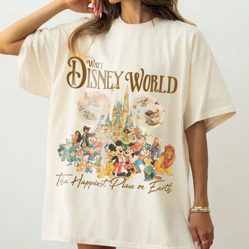 May include: Cream-colored t-shirt featuring the text "Walt Disney World" in brown script above a colorful graphic of Disney characters and Cinderella's castle. The phrase "The Happiest Place on Earth" is printed below the graphic.