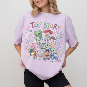 May include: A light purple t-shirt featuring the words "Toy Story" in colorful letters, along with cartoon illustrations of characters from the movie. The design includes Woody, Buzz Lightyear, and Rex.