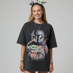 May include: Dark gray t-shirt featuring a graphic of The Mandalorian and Grogu. The design includes the Mandalorian's helmet and Grogu. The person is wearing a headband with Grogu and a silver ball on it.
