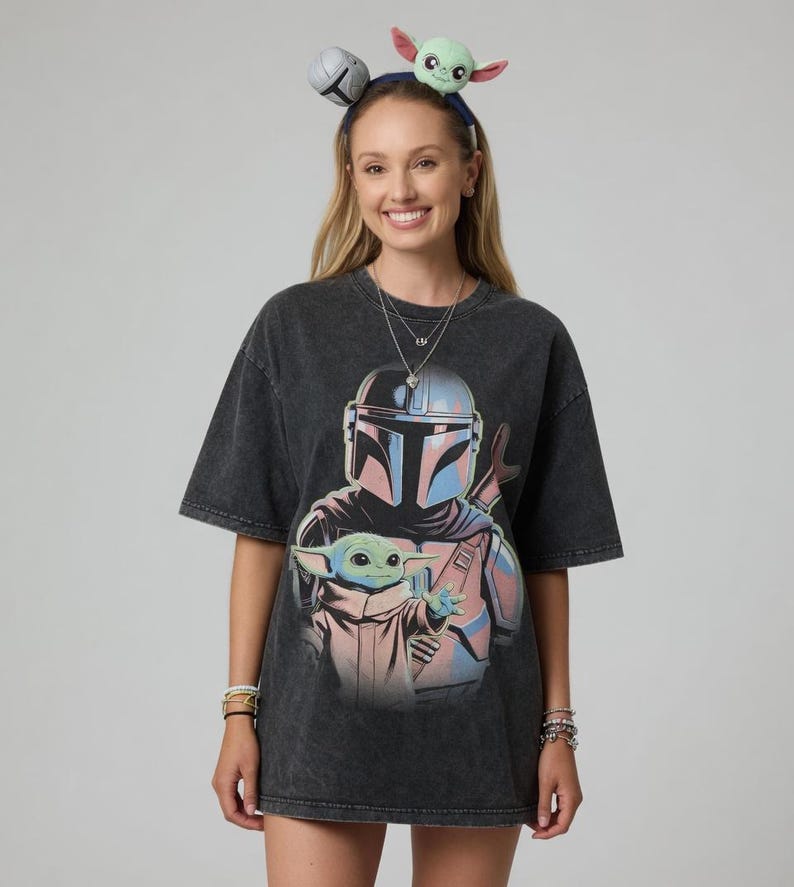 May include: Dark gray t-shirt featuring a graphic of The Mandalorian and Grogu. The design includes the Mandalorian's helmet and Grogu. The person is wearing a headband with Grogu and a silver ball on it.