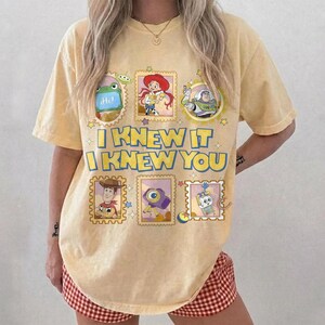 May include: A pale yellow short-sleeve t-shirt featuring a graphic print of Toy Story characters in framed boxes. The text "I KNEW IT I KNEW YOU" is printed in large blue letters in the center, surrounded by small stars and colorful icons.