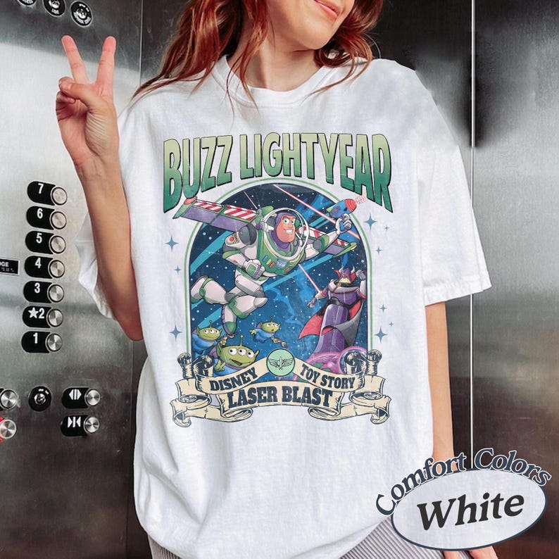 May include: A white t-shirt featuring a graphic print of Buzz Lightyear and Zurg in space. The text reads BUZZ LIGHTYEAR at the top and DISNEY TOY STORY LASER BLAST at the bottom. The design includes green, purple, and blue colors with stars.