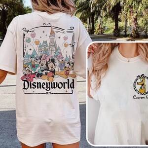May include: Cream-colored t-shirt featuring a Disneyworld graphic on the back with Mickey, Minnie, Donald, Daisy, and Goofy. The front has a Pluto graphic inside a checkered circle. The text "Disneyworld" and "1971" are also visible.