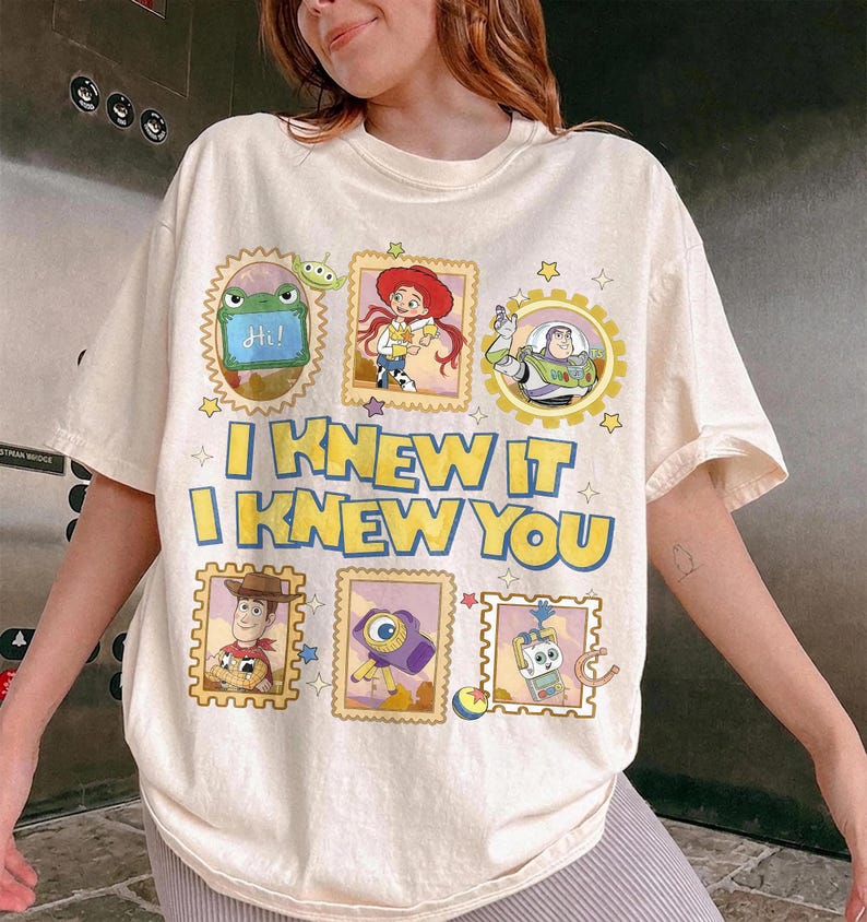 May include: A cream-colored t-shirt featuring a graphic print of various Toy Story characters in postage stamp-style frames. The text "I KNEW IT I KNEW YOU" is printed in large yellow letters with blue outlines across the center of the shirt.