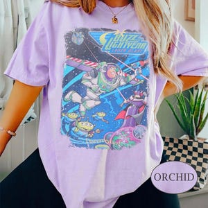 May include: A light purple t-shirt featuring a graphic print of Buzz Lightyear from Laser Blast. The design includes Buzz Lightyear, green aliens, and Zurg in a space setting with the text Buzz Lightyear Laser Blast and Disneyland.