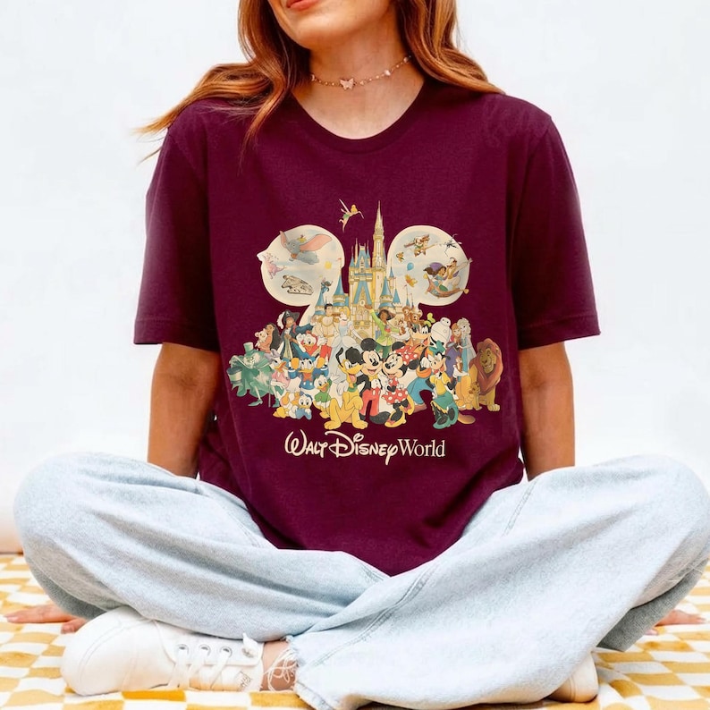 May include: Burgundy t-shirt featuring a colorful graphic of Disney characters and the Walt Disney World logo. The design includes Mickey Mouse, Donald Duck, and other beloved characters. The shirt has short sleeves and a crew neck.