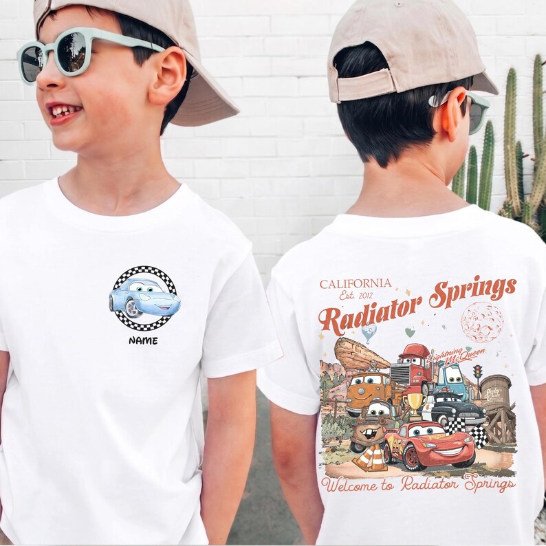 Puede incluir: Camisetas blancas con dise&ntilde;os de coches de dibujos animados. Una camiseta tiene un gr&aacute;fico frontal de un coche azul dentro de un c&iacute;rculo a cuadros con la palabra "NAME" debajo. La otra camiseta tiene un gr&aacute;fico trasero con el texto "CALIFORNIA Est. 2012 Radiator Springs".