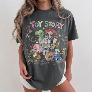 May include: Dark gray t-shirt featuring the words "Toy Story" in colorful lettering, along with cartoon characters like Woody, Buzz Lightyear, and Rex. The design has a hand-drawn aesthetic with scribbled lines.