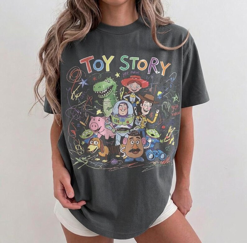 May include: Dark gray t-shirt featuring the words "Toy Story" in colorful lettering, along with cartoon characters like Woody, Buzz Lightyear, and Rex. The design has a hand-drawn aesthetic with scribbled lines.